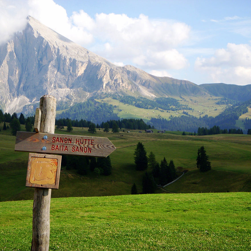 Wooden signpost for walkers in Ortisei, Italy. The weather is sunny and there is green grass, trees and mountains in the background.